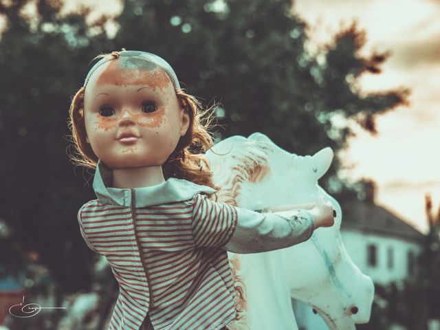 A worn vintage doll in a striped dress riding a pale carousel horse outdoors