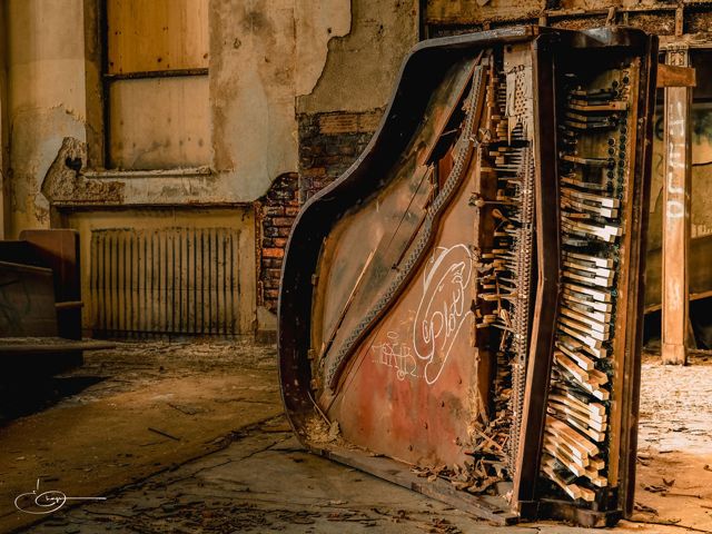 An abandoned upright piano with its front torn away, keys and strings exposed, in a decaying room