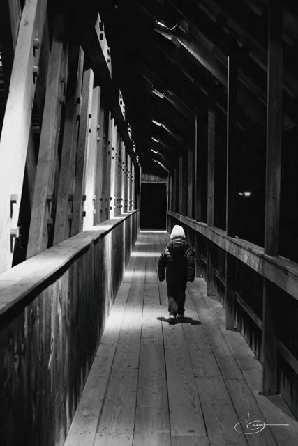 Black and white view down the inside of a long wooden covered bridge, with one person walking toward the far end