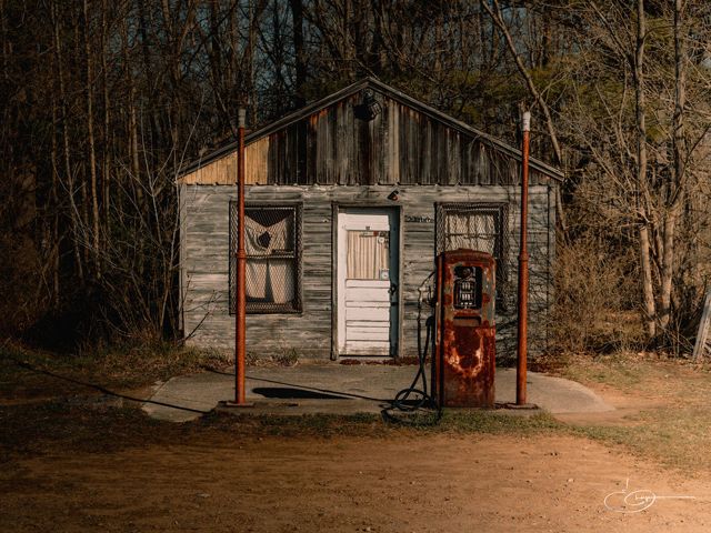 A weathered wooden shack with a rusted vintage gas pump out front, at the edge of a bare autumn woods