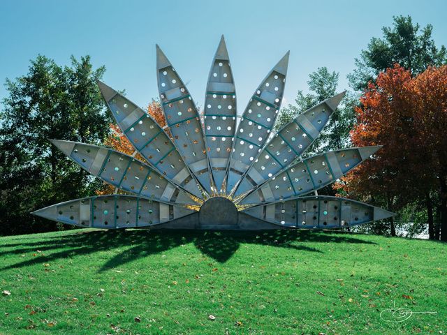 A large metal sunburst sculpture with pointed rays standing on a green lawn, autumn trees behind it