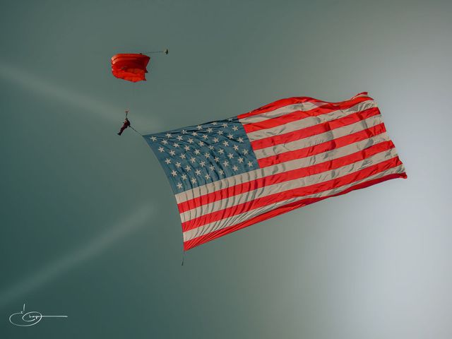 A skydiver under a red parachute trailing a large American flag across a pale sky