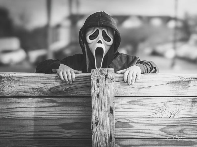 Black and white photo of a child in a Ghostface Halloween mask peeking over a wooden fence