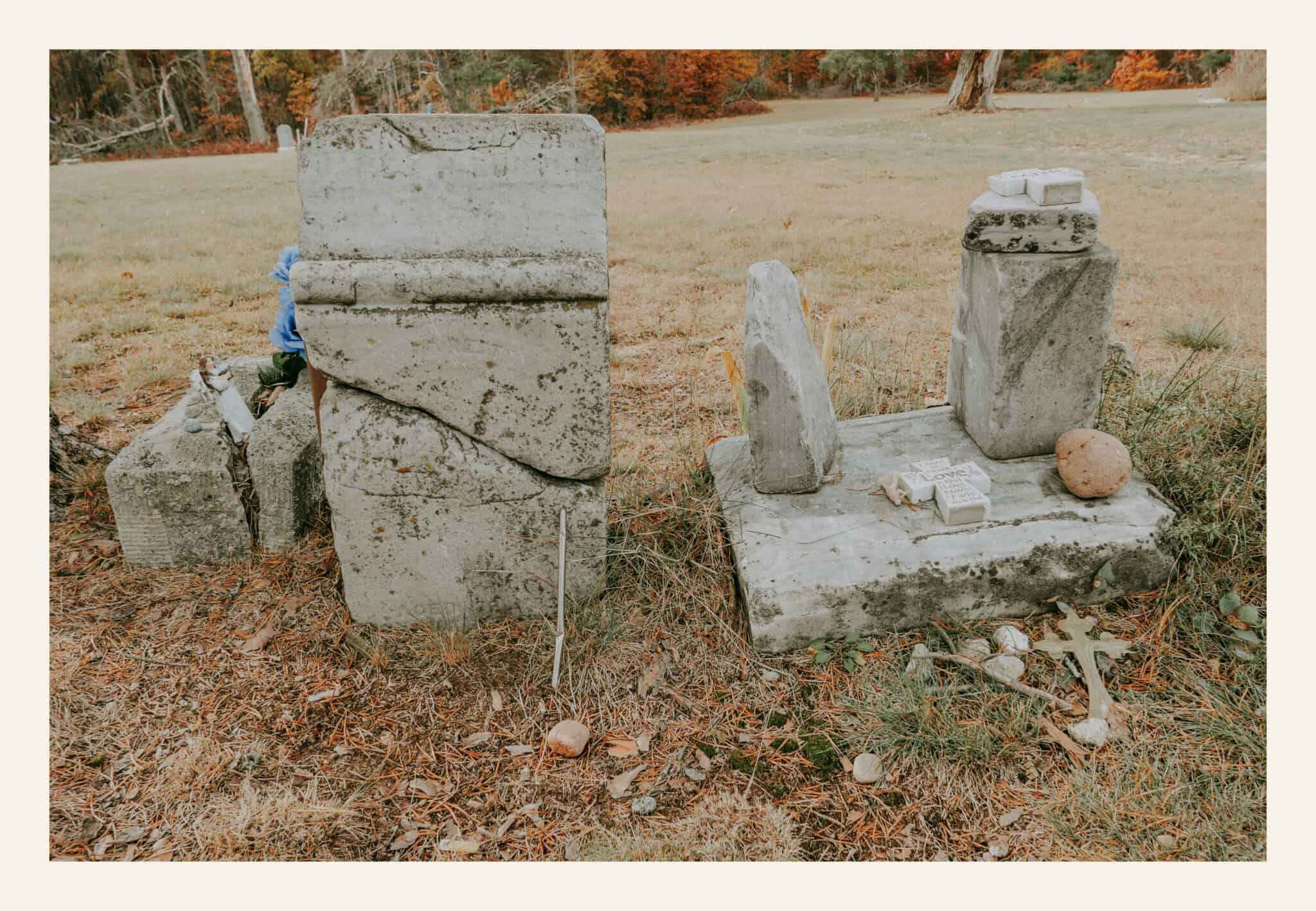 Broken limestone headstones at Pere Cheney Cemetery, Michigan, surrounded by pine needles and faded blue artificial flowers. The cracked stones show the age and neglect of this historic ghost town graveyard.